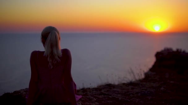 Seorang gadis melihat matahari terbenam di pantai laut di atas batu duduk — Stok Video