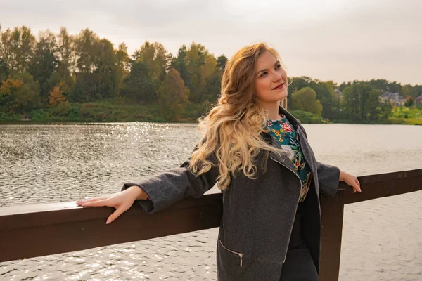 Chica alegre De pie cerca del estanque con labios tiernos con una sonrisa perfecta en un abrigo en el otoño en el muelle por la tarde — Foto de Stock