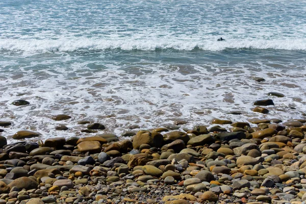 遠くで波を戦う孤独なスイマーと岩のビーチでさわやかな寒さ — ストック写真