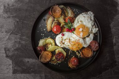 Kızarmış yumurtaları, ızgara domatesleri, biberleri, havuçları ve patatesleri siyah tabakta, tahtadan yapılmış sağlıklı kahvaltı ve ev yapımı pişirme konseptiyle kapat.