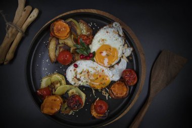 Kızarmış yumurtaları, ızgara domatesleri, biberleri, havuçları ve patatesleri siyah tabakta, tahtadan yapılmış sağlıklı kahvaltı ve ev yapımı pişirme konseptiyle kapat.