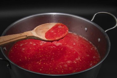 Ahşap kaşıkta domates sosu siyah arka planda metal tencerenin üzerinde taze sebzeler ve organik gıdalar için ev yapımı pişirme hazırlığında tutulur.