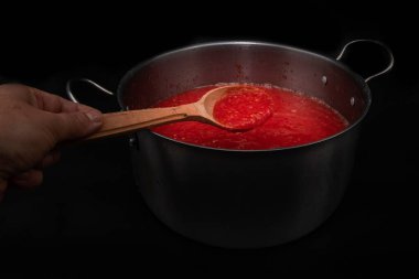 Ahşap kaşıkta domates sosu siyah arka planda metal tencerenin üzerinde taze sebzeler ve organik gıdalar için ev yapımı pişirme hazırlığında tutulur.