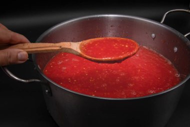 Ahşap kaşıkta domates sosu siyah arka planda metal tencerenin üzerinde taze sebzeler ve organik gıdalar için ev yapımı pişirme hazırlığında tutulur.