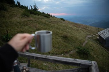 El ele tutuşan metal kamp kupası ve dağ manzarası. Arka planda seyahat macerası. Sabah kahvesi, açık hava yaşam tarzı ve kamp yapma kavramı.