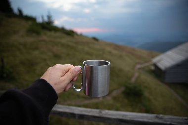 El ele tutuşan metal kamp kupası ve dağ manzarası. Arka planda seyahat macerası. Sabah kahvesi, açık hava yaşam tarzı ve kamp yapma kavramı.