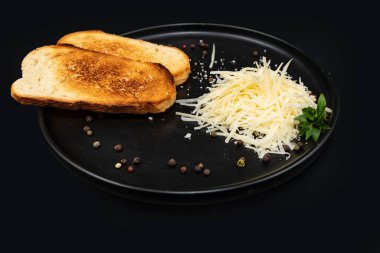 Kızarmış ekmek ve rendelenmiş peynir, siyah bir tabakta karabiber ve taze otla servis ediliyor. Minimalist yemek tasarımı, yemek pişirme, kahvaltı, yemek tarifleri ve restoran menüleri için mükemmel..