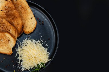 Kızarmış ekmek ve rendelenmiş peynir, siyah bir tabakta karabiber ve taze otla servis ediliyor. Minimalist yemek tasarımı, yemek pişirme, kahvaltı, yemek tarifleri ve restoran menüleri için mükemmel..