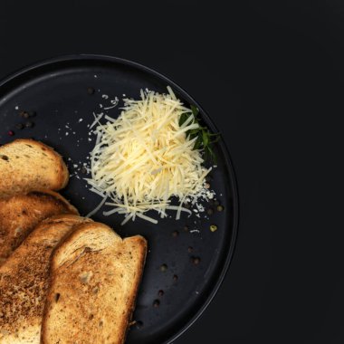 Kızarmış ekmek ve rendelenmiş peynir, siyah bir tabakta karabiber ve taze otla servis ediliyor. Minimalist yemek tasarımı, yemek pişirme, kahvaltı, yemek tarifleri ve restoran menüleri için mükemmel..