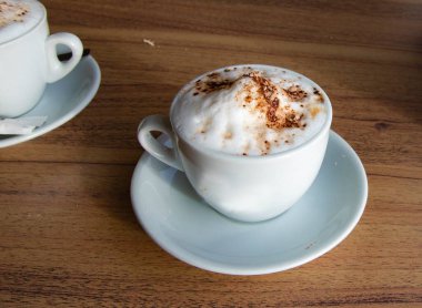 Kapuçinoyu kapuçino üzerine süt köpüğü ve tarçın tozu serpiştirilmiş beyaz bir bardakta servis et. Sabah kahvesi, rahat kafe atmosferi ve yaşam tarzı kavramı..