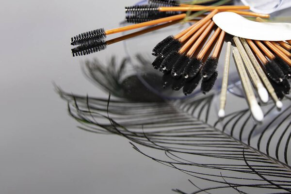 Закрыть павлинье перо, помещенное набором одноразовых щёток для ресниц на отражающую поверхность. Концепция красоты, косметики, креативности и профессиональных средств макияжа.