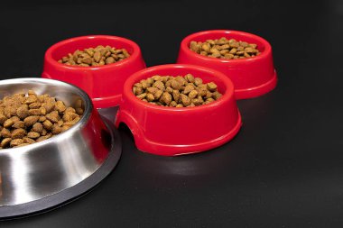 Üç kırmızı plastik kasede servis edilen kuru evcil hayvan yiyeceği ve siyah arka planda paslanmaz çelik kase. Hayvan bakımı, beslenme ve beslenme araçları kavramı.