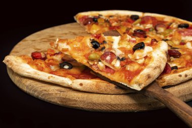 Eritilmiş peynirli, zeytinli, sosisli ve sebzeli sıcak pizza dilimini ahşap bir tahtaya yakın. Restoran menüsü, yemek servisi, İtalyan mutfağı ve fast food konseptleri için mükemmel..