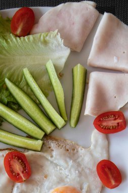 Kızarmış yumurta, salatalık çubuğu, marul, vişneli domates ve beyaz tabakta servis edilen jambonlu sağlıklı bir kahvaltı. Besleyici yemek ve diyet gıda konsepti.