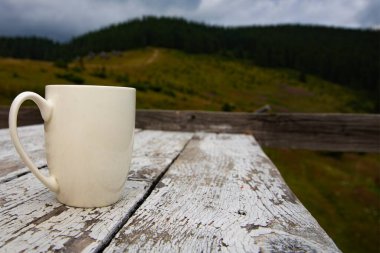 Manzaralı dağ manzarasına karşı eski ahşap masanın üzerinde beyaz seramik bardak. Seyahat, rahatlama, sabah kahvesi ve kırsal yaşam tarzı kavramı.