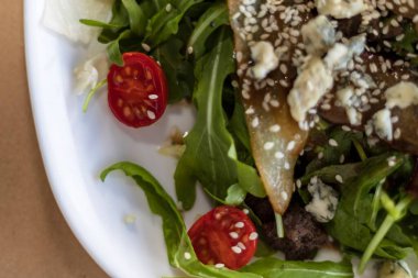 Taze roka, kavrulmuş armut dilimleri, mavi peynir parçaları, vişneli domatesler ve susamlı tohumlardan oluşan, beyaz bir tabakta servis edilen gurme salatasının yakın çekimi. Sağlıklı yemek, restoran yemeği ve diyet konsepti..