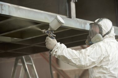 Beyaz üniformalı bir adam metal bir ürüne sprey tabancası ile boya uygular..