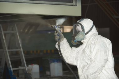 Beyaz üniformalı bir adam metal bir ürüne sprey tabancası ile boya uygular..