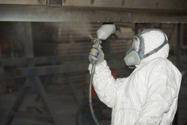 Beyaz üniformalı bir adam metal bir ürüne sprey tabancası ile boya uygular..