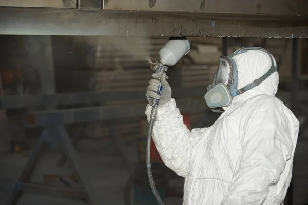 Um homem de uniforme branco aplica tinta com uma pistola de pulverização em um produto de metal . — Fotografia de Stock