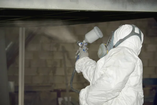 Beyaz üniformalı bir adam metal bir ürüne sprey tabancası ile boya uygular..