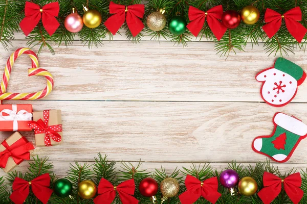 Рождественская рама из еловых ветвей, украшенных шарами, луками, конфетами и коробками на светлом деревянном фоне — стоковое фото
