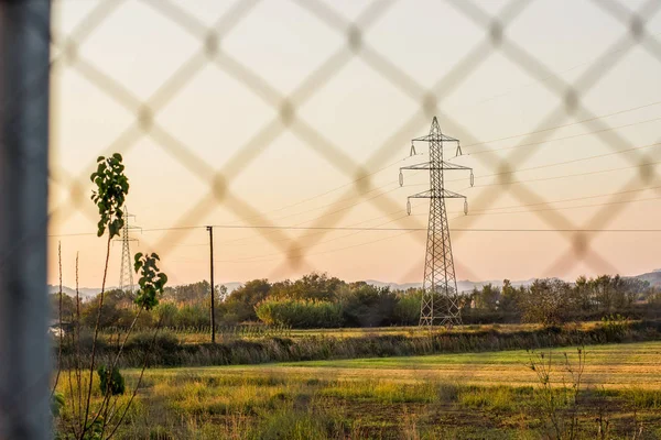 high voltage tower industry infrastructure object in rural country side field environment through iron grid fence in soft focus evening calming time