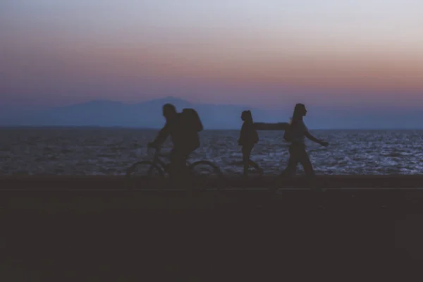 creative unfocused humans wallpaper pattern of fuzzy walking people silhouettes on dark twilight waterfront along Pacific ocean and unfocused mountain horizon background in evening sunset time