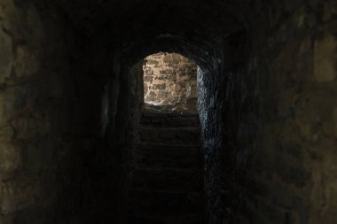 Ortaçağ yeraltı kalesinde alacakaranlık aydınlığı soğuk taş duvarlar ve çıkış manzarası arasındaki dar geçit. 