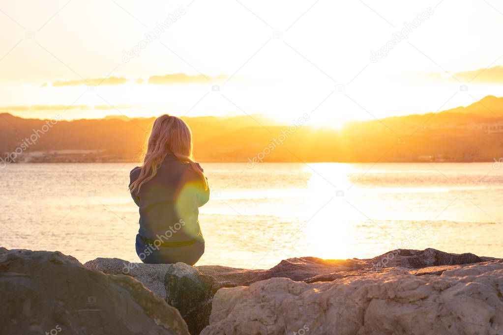 loneliness feelings of sitting girl on stone shore line near sea waterfront in bright orange sunset time with glares moody photography poster with empty copy space for text