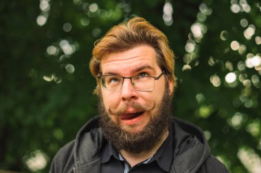 Komik bir portre, suratını buruşturan orta yaşlı sakallı bir adam park alanının dışında gözlüklü, odaklanmamış bir bokeh arka plan manzaralı.