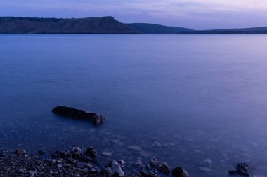 Manzaraya uzun süre maruz kalan nehir kıyısı çizgisi, yumuşak odak taşıyla sığ sularda ön planda ve tepenin arka planında alacakaranlık aydınlığında alacakaranlık aydınlığında ufuk çizgisi.
