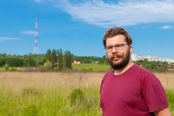 middle adult man portrait with beard and glasses on summer time bright clear weather landscape outskirts scenic view with rustic buildings and TV tower copy