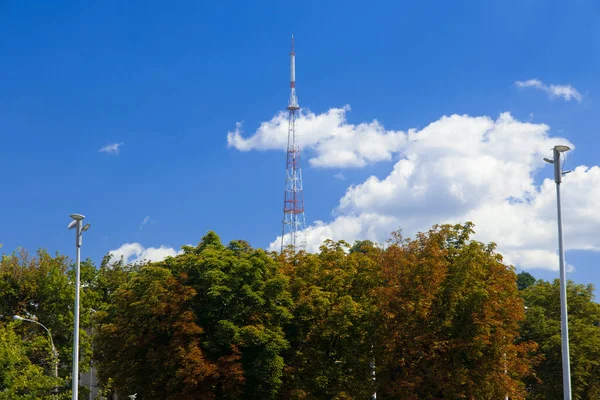 TV tower communication infrastructure object on top of hill in park outdoor district with a lot green foliage trees and street lantern foreground in clear weather day time