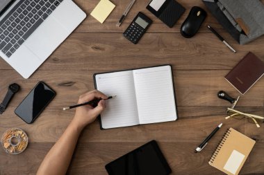 Boş dizüstü bilgisayar listesinde sol el kalem, düz masa üzerinde ofis aksesuarları. Liste yapmak ve dizüstü bilgisayar konseptinde hatırlatma listesi yapmak için ofis masası düzleminde çalışıyorum..