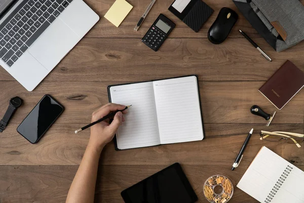 La mano izquierda que sostiene el lápiz en la lista vacía del cuaderno con los accesorios de oficina en la disposición plana de la mesa.Trabajar en la disposición plana del escritorio de la oficina.Para hacer la lista y la lista del recordatorio de la reunión en concepto del cuaderno . — Foto de Stock