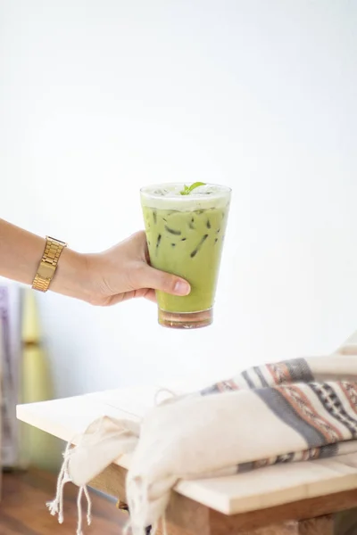 Teh hijau es atau matcha latte pada gelas tinggi dengan jerami di meja kayu putih dan dekorasi pakaian object.Hand memegang gelas minuman . — Stok Foto