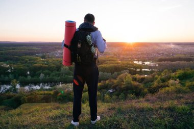 Bir tepenin üstünde sırt çantalı genç bir turist akşam üzeri ufka ve manzaraya bakıyor..