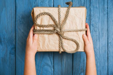 Elinde hediye tutan bir çocuğun elleri, mavi ahşap arka planda üst manzara.