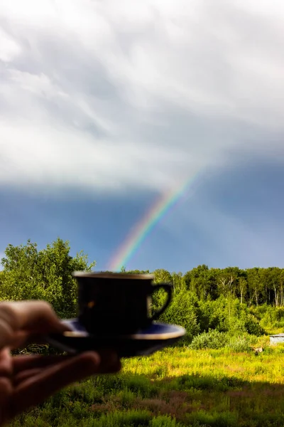 Rainbow over the forest. Mug of coffee, early morning, cloudy sky and clear colors of the rainbow, forest road. Natural landscape. Rainbow colors after rain. Rain clouds.