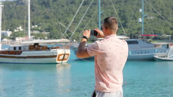 Молодой человек делает панорамное фото или видео со смартфоном возле морского пляжа — стоковое видео