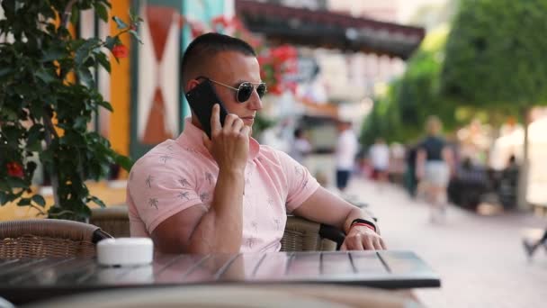 Νεαρός άνδρας μιλάει με smartphone που κάθεται στο υπαίθριο καφέ — Αρχείο Βίντεο