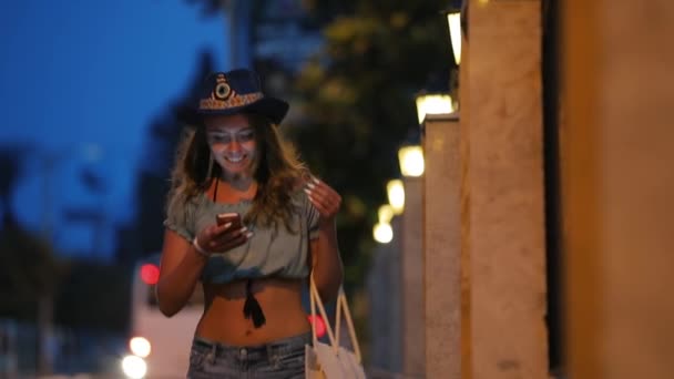 Mujer feliz en el uso de sombrero smartphone en la noche calle de la ciudad — Vídeos de Stock