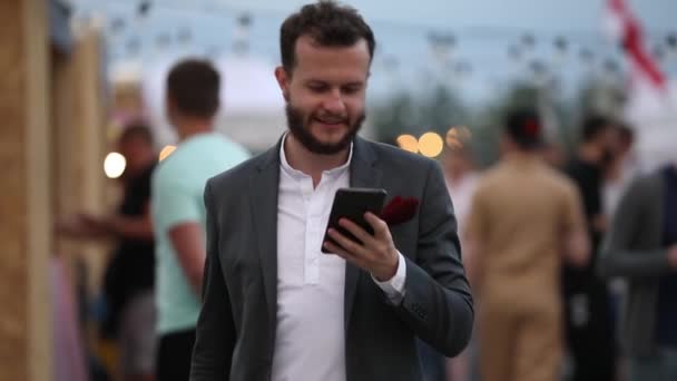 Ευτυχισμένος γενειοφόρος άνδρας σε κλασσικό κοστούμι περπάτημα ψάχνει για smartphone — Αρχείο Βίντεο