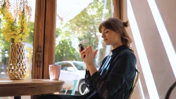 Щаслива дівчина, що розслабляється в кафе, сидить біля вікна, використовує смартфон — стокове відео
