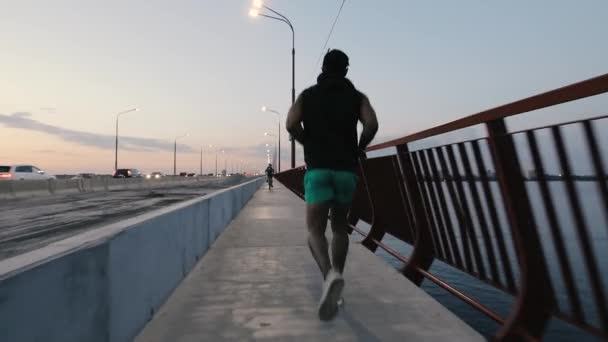 Man joggen op een brug in de stad op de avond, achteraanzicht — Stockvideo