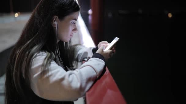 Wanita muda menggunakan aplikasi smartphone berdiri di jembatan pada waktu malam — Stok Video