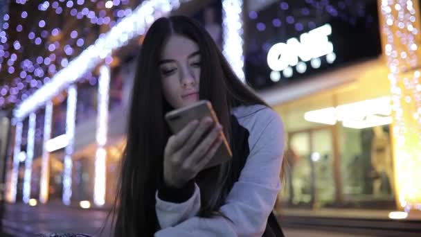 Женщина использует смартфон, сидя в центре города ночью — стоковое видео