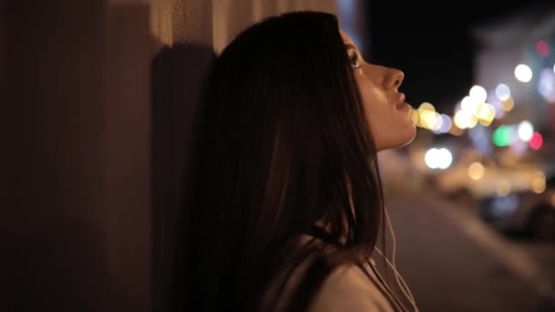 Жінка слухає музику з навушниками і розслабляється, спираючись на стіну в нічному місті — стокове відео