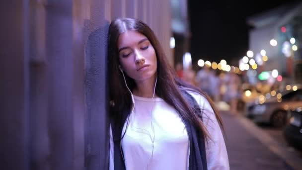 Mujer escuchando música y relajándose apoyada en la pared de la ciudad nocturna — Vídeos de Stock